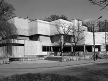 <p style="text-align: justify;">The Kaunas Picture Gallery, opened in 1979 to house the artworks of famous Lithuanian collector Mykolas Žilinskas, is a striking example of architecture of the period, combining elements of Brutalism and sleek International Style modernism.</p>
<p style="text-align: justify;">The design of the gallery emphasises the built form as sculpture; elevated and distinguished from the street by a stepped plinth, which serves as a podium presenting the modelled architecture, just as artworks are exhibited within the building. The sculptural quality of the massed cuboid volumes is further enhanced by the scarcity of windows on the upper floors, displaying a smooth concrete surface which stresses form over decorative elements. Such a dramatic lack of windows, and attendance to massive sculptural blocking, is reminiscent of parts of Claude Paillard’s Theater St Gallen, built in the 60s. Use of glass, and permeable elements is restricted to the the first floor. Here, the large curtains windows and open terrace produce an interruption in the overall sculptural solidity of the gallery building. This serves to definitively accent the composition above, and create a diagonal line symmetry between the stepped forms of the plinth, and those of the building above. The composition itself utilises a strict geometric scheme, made up of cuboid volumes held together in a dynamic arrangement (even the piloti are square-forms). Generally a horizontal composition, which welcomes a panoramic view, this is broken by the light-handed gesture of chimney-like vertical forms that rise above the entrance, and provide a signpost of this in what is generally a highly non hierarchical arrangement. The entrance to the museum today is further highlighted by Jadvyga Mozuritė-Klemkienė’s sculpture <em>Creator</em>, made in 1987, and in 2007, one of the gallery’s corner walls was decorated by a work of the Brazilian street artist team Os Gemeos.</p>
<p style="text-align: justify;">While much of the scheme of this building accentuates its architectural exceptionality, the Kaunas Picture Gallery is also artfully integrated into its site on the curve of K. Donelaičio street and backing onto the slope of Žaliakalnis. The irregularly stepped plinth that leads to the entrance of the gallery integrates this approach into the hill behind. Steps ascending this plinth from different angles off the street create an open transition from the pavement, attentive to the corner that the building sits upon. This corner itself finds itself represented in the composition of the structure; the two offset levels of repeated stepping on the left hand side of the gallery may be read as a formalisation of this corner. Finally, the terrace space created by the overhang on the right-hand side of the gallery, and held in by the square piloti, softens the divide between environment and walled interior in this building, through a semi-contained, transitional space.</p>
<p style="text-align: justify;">Decoration in this building is mainly restricted to the interior, and delicate, or hidden gestures on the outside surfaces. Diagonal stone striations in the surfaces of the plinth texturally distinguish it from the building proper (and perhaps blend it with the “natural”hill behind). Elsewhere, the roof of the terrace is clad with geometric relief tiles, and a band of dark stone wraps around the corner at street level. A slight lip at the roofline, and windows, further draws out the sculptural form of this building.</p>
<p style="text-align: justify;">The interior decoration of the gallery employs a range of materials fashionable in the period: dolomite, light granite stucco, and dark granite slabs. The interior of the gallery is arranged around the main staircase. On the first floor stands the main hall- used for temporary exhibitions-, a lobby, hallway and cafe. The second and third floors are made up of exhibition space, as well as an auditorium for special events. Formerly, an artist’s studio was housed here. Up to the day, the lobby’s authentic interior has been preserved, decorated with original furniture, light fixtures, and wooden finish.</p>
<p style="text-align: justify;"><em>Ellen Tracy</em></p>